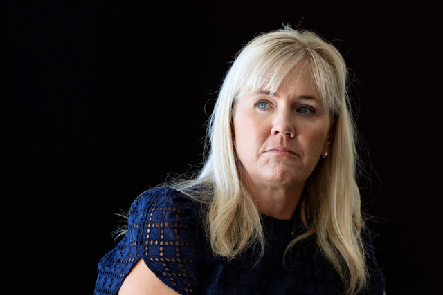 A blonde woman with light skin and blue eyes wears a navy blue top with a lace pattern, looking to her right with a serious expression against a black background.